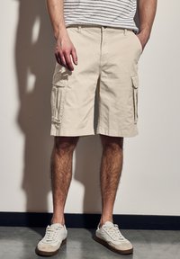 Béžové cargo šortky se bočními kapsami, vyrobené z lehké látky. Nosené s bílými teniskami, mají volný střih a délku nad kolena.