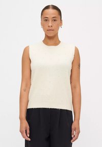 Mouwloze crèmekleurige trui met een gestructureerde afwerking, voorzien van verhoogde bobbel-details en een ribgebreide halslijn, gecombineerd met donkere broeken.
