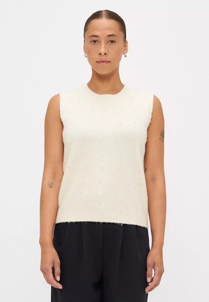 Maglione senza maniche color crema con una finitura testurizzata, caratterizzato da dettagli a pallini in rilievo e un colletto a coste, abbinato a pantaloni scuri.