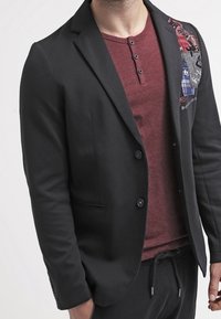 Blazer nero con un design aderente, caratterizzato da un colletto strutturato e un accento di patch colorato. Indossato sopra una maglietta henley bordeaux.