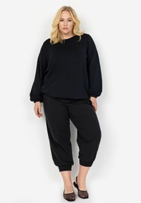 Fekete, hosszú ujjú felső, gumis mandzsettákkal és fekete, tapered fazonú nadrág, gumis szegéllyel. A modellen fekete hálós balerina cipők vannak. Egyszerű, hétköznapi dizájn.