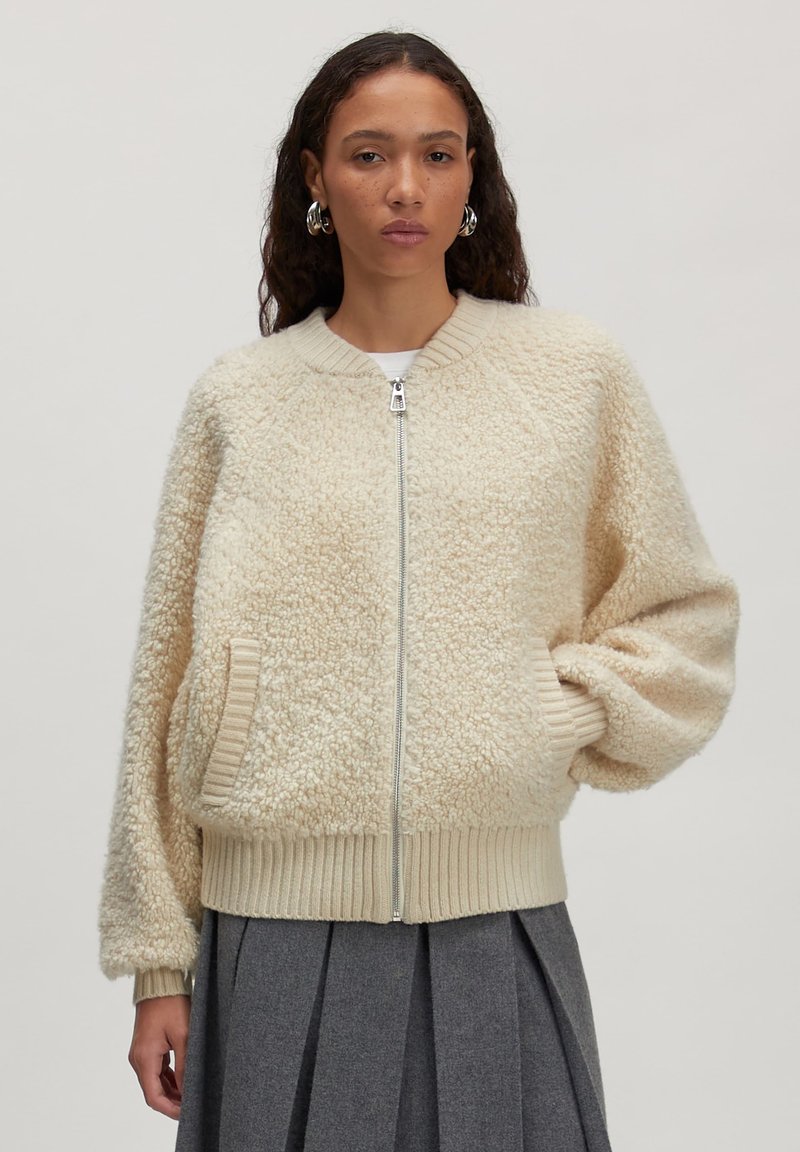 Beige bomberjack gemaakt van gestructureerde fleece, met een voorrits, ribgebreide manchetten en zijzakken. Gecombineerd met een geplooide grijze rok.