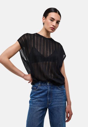 Femme aux cheveux foncés portant un haut noir transparent à rayures sur un soutien-gorge noir, assorti à un jean bleu taille haute, posant devant un fond uni.