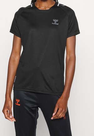 Persona con camiseta deportiva negra de manga corta y pantalones con logotipos grises y rojos de Hummel sobre un fondo claro y sencillo.