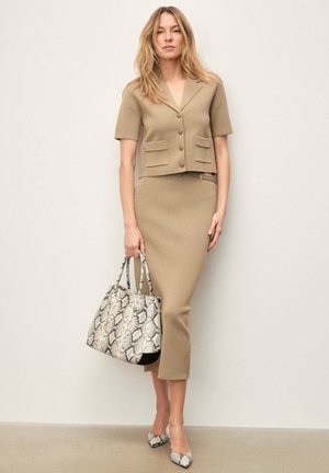 Ženska, oblečena v bež jakno s kratkimi rokavi z gumbi in ujemajočo midi krilo, drži torbo z vzorcem kačje kože in nosi usklajene pete.