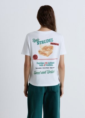 Persona che indossa una maglietta bianca con una grafica di strudel di mele e testo sulla schiena, abbinata a pantaloni verde scuro.