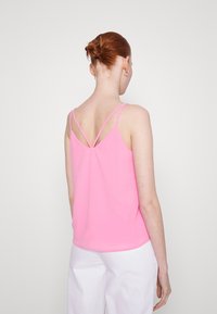 Pink camisole with thin straps, featuring a crisscross back design. Smooth fabric; slightly loose fit, paired with white trousers.