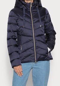 Veste matelassée bleu marine avec capuche, fermeture éclair, accents dorés et coutures en chevrons. Comprend des poches latérales et des panneaux élastiques sur les côtés pour un ajustement parfait.
