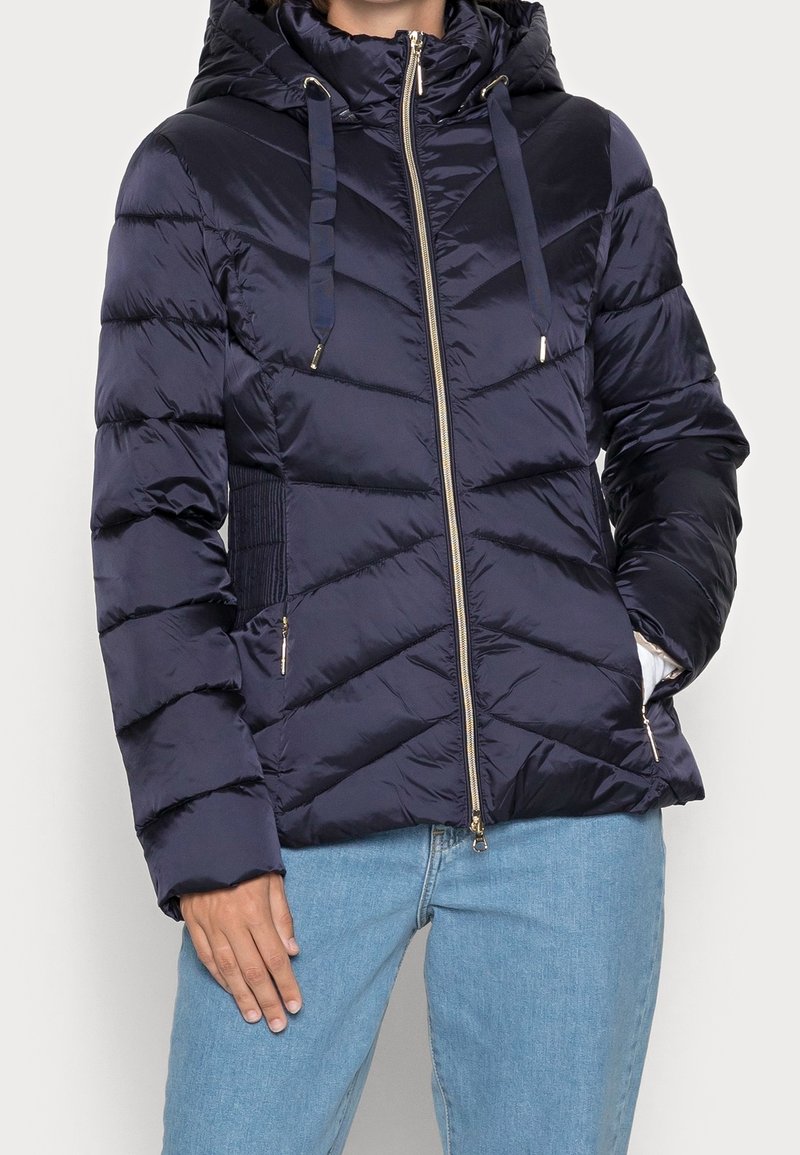 Veste matelassée bleu marine avec capuche, fermeture éclair, accents dorés et coutures en chevrons. Comprend des poches latérales et des panneaux élastiques sur les côtés pour un ajustement parfait.