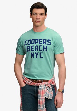 Mann trägt ein mintgrünes T-Shirt mit dem Text "COOPERS BEACH NYC", dunkle Jeans, ein kariertes Hemd um die Taille gebunden und eine Armbanduhr.