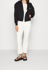 Černá mikina na zip s kapucí, pod ní bílý tričko, bílé rozšířené džíny a černé sandály na podpatku s tlustou podrážkou.