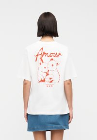 Γυναίκα με κοντά σκούρα μαλλιά που φοράει λευκό μπλουζάκι με κόκκινη ζωγραφιά ζώου και κείμενο «Amour», σε συνδυασμό με τζιν φούστα.