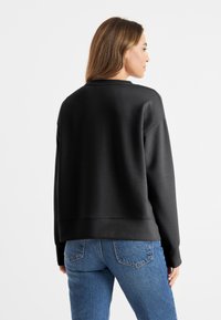 Zwarte sweatshirt met een ontspannen pasvorm, geribbelde manchetten en onderrand, zachte textuur, gecombineerd met hoog-getailleerde blauwe jeans. Achteraanzicht.