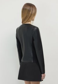 Giacca di pelle nera con un design aderente e maniche lunghe, caratterizzata da una texture liscia e un colletto rotondo, abbinata a una gonna nera.