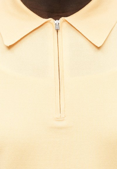 Μπεζ πλεκτό πόλο μπλουζάκι με διπλωμένο γιακά και μικρό ασημί φερμουάρ στο μπροστινό κέντρο του λαιμού.