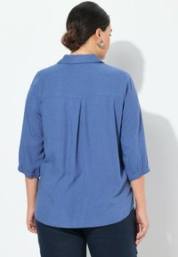 Camicia blu con bottoni realizzata in tessuto leggero, con maniche a tre quarti, polsini piegati, dettaglio di piega sul retro e orlo curvo.
