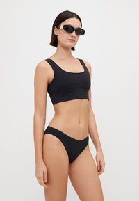 Fekete, két részes bikini, amelynek a felső része négyzet alakú nyakkivágással és alacsony derekú alsó résszel rendelkezik. Sima textúra minimális varratokkal és díszítésekkel.