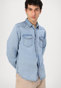 Camisa de mezclilla azul claro con mangas largas, dos bolsillos en el pecho y un cierre de botones en la parte delantera. Presenta un cuello clásico y un dobladillo ligeramente curvado.
