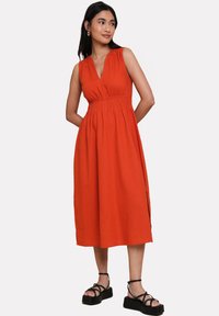 Vestido naranja sin mangas con escote en V y cintura fruncida. De longitud media con aberturas laterales, combinado con sandalias de plataforma negras con tiras.