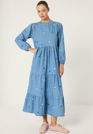 Γυναίκα με μακρύ μπλε φόρεμα με πολύχρωμα μικρά λουλουδάτα μοτίβα και μακριά μανίκια, σε συνδυασμό με ροζ αθλητικά παπούτσια, στέκεται μπροστά σε λευκό φόντο.