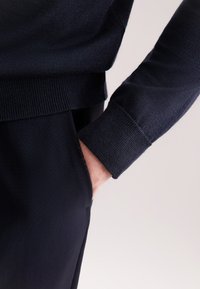 Persona che indossa un maglione blu scuro e pantaloni abbinati, con la mano destra comodamente infilata in tasca.