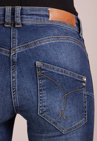 Vaqueros de mezclilla azul oscuro con un diseño ajustado, bolsa trasera prominente con costura amarilla y una etiqueta de cuero en la cintura.