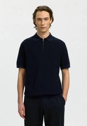 Mężczyzna o średniej długości włosach, ubrany w granatowy polo z krótkim rękawem na suwak oraz czarne spodnie, stojący na jasnym, jednolitym tle.