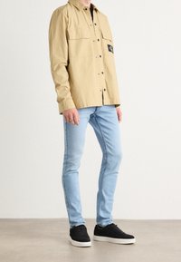 Camisa beige de manga larga con dos bolsillos en el pecho y parche negro, combinada con jeans ajustados azul claro y zapatillas negras.