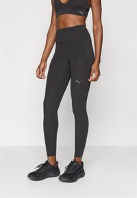 Kobieta ubrana w czarne sportowe legginsy z wysokim stanem i dopasowany czarny sportowy stanik z logo Puma, w połączeniu z czarnymi butami do biegania Puma.