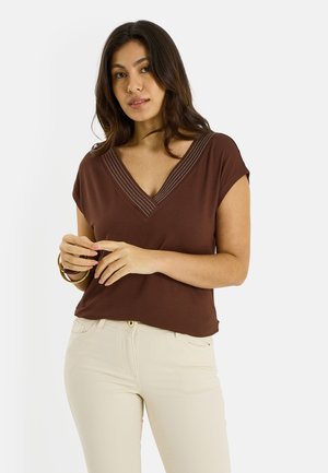 Mujer de cabello largo y oscuro, vestida con una blusa marrón de cuello en V y pantalones beige, con las manos juntas cerca de la cintura y pulseras doradas en la muñeca.