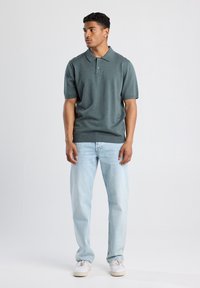 Camisa polo verde de manga corta con cuello acanalado, combinada con jeans azul claro y zapatillas blancas, de pie contra un fondo neutral.