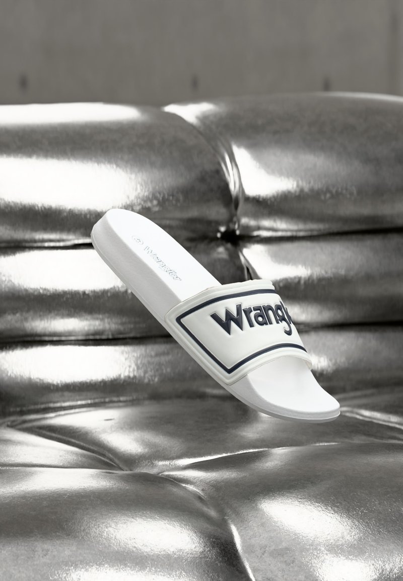 Λευκό σανδάλι slide με το λογότυπο "Wrangler" στο λουράκι, ακουμπισμένο σε γυαλιστερή ασημένια μεταλλική επιφάνεια με καπιτονέ μοτίβο.
