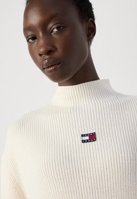 Cream ribbed knit sweater with a high collar and a small red, white, and blue "TOMMY JEANS" logo patch on the chest area.
