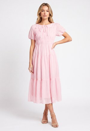 Femme aux longs cheveux blonds portant une robe midi rose clair à manches courtes, évasée par étages, et des escarpins beiges à bride cheville, debout avec une main sur la hanche.