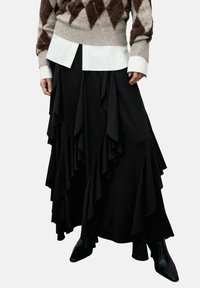 Suéter con patrón de diamantes marrón y beige, camisa blanca debajo, falda negra con volantes superpuestos y botas de punta negra.