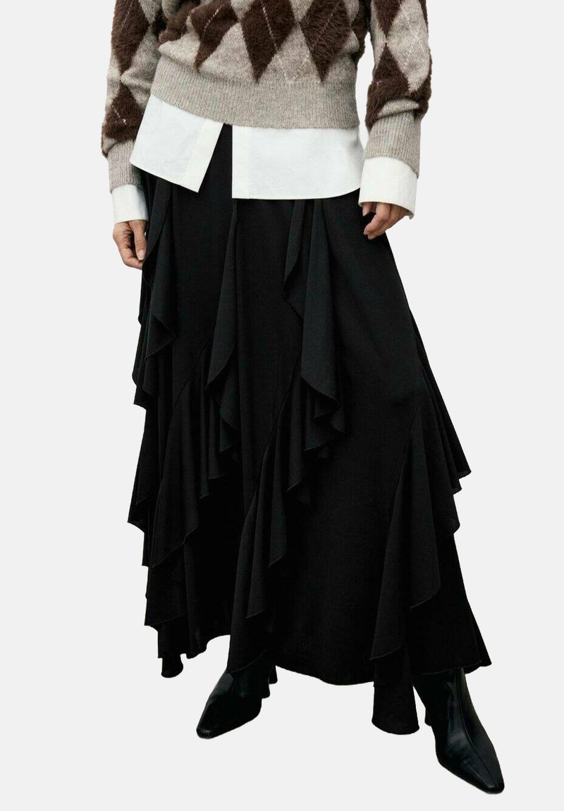 Suéter con patrón de diamantes marrón y beige, camisa blanca debajo, falda negra con volantes superpuestos y botas de punta negra.