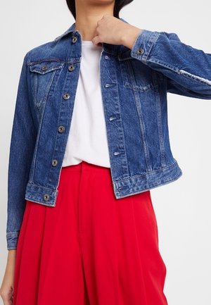 Personne portant une veste en jean bleue sur une chemise blanche et un pantalon rouge taille haute, ajustant le col de la veste d'une main.
