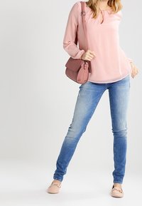 Blouse rose ample avec un détail en trou de serrure, jean skinny bleu avec léger vieillissement, et ballerines rose clair, complété par un sac bandoulière assorti.