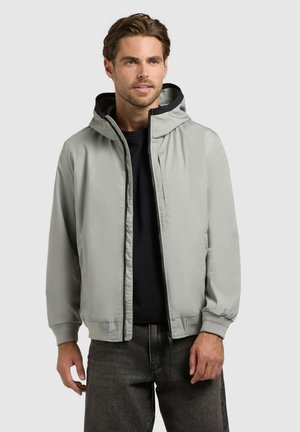 Man draagt een lichtgrijs gewatteerd jack over een zwart shirt en donkergrijze spijkerbroek, staand tegen een effen lichte achtergrond.