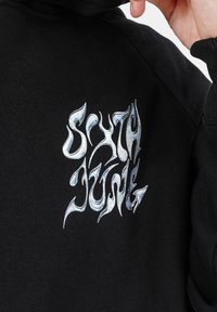 Sudadera negra con un diseño bordado en plata metálica en la parte posterior que muestra el texto "SIXTH JUNE" en una fuente con forma de llama.