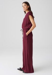 Burgunderfarbener ärmelloser Jumpsuit mit einer taillierten Taille, Seitentaschen und einem lockeren, fließenden Schnitt. Weicher Stoff mit glatter Textur.