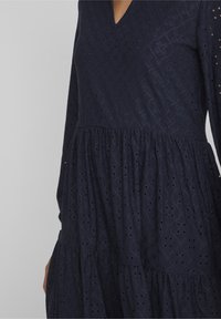 Marinblå klänning med V-halsringning, långa ärmar och en flera lager-skirt. Har ett strukturmönster med hålmönster och ribbade detaljer.