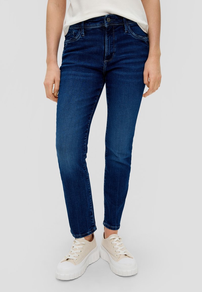 Jean en denim bleu foncé à coupe ajustée, présentant cinq poches, passants pour ceinture et une légère décoloration subtile. Associé à des baskets à plateforme beige.