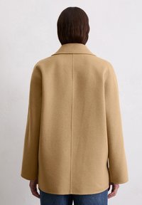 Beige wool coat with a relaxed shape, large collar, and dropped shoulders. Center seam and long sleeves, styled with jeans.