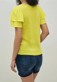 Gul kortærmet top med lagdelte pufærmer og en tekstureret strik, parret med mørke denimshorts med baglommer.