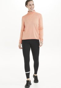 Letvægts, langærmet top med lynlås i koral, med en struktureret overflade. Båret sammen med sorte leggings med reflekterende detaljer.