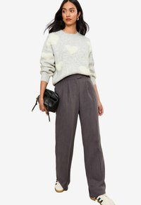Pull gris avec des motifs de cœurs blancs duveteux, assorti à un pantalon plissé gris foncé taille haute. Tient un petit sac noir, porte des baskets.