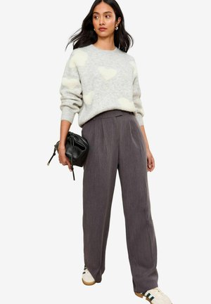 Maglione grigio con motivi a cuore bianchi pelosi, abbinato a pantaloni scuri grigi a vita alta e plissettati. Tiene in mano una piccola borsa nera e indossa delle sneakers.