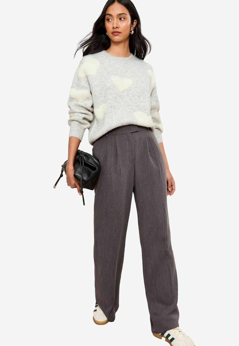 Pull gris avec des motifs de cœurs blancs duveteux, assorti à un pantalon plissé gris foncé taille haute. Tient un petit sac noir, porte des baskets.