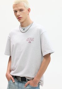Grå kortærmet t-shirt med broderet "STUD" logo i rødt. Har en afslappet pasform, rund hals, og er parret med lyseblå denim.